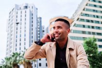 Baixo ângulo de positivo bem vestido jovem empresário hispânico falando no smartphone e discutindo notícias na rua urbana com edifícios contemporâneos em segundo plano — Fotografia de Stock