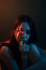 Jeune modèle féminin sans émotion avec projection de lumière en forme de croix sur le visage assis dans un studio sombre et regardant loin — Photo de stock