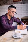 Side view of handsome young Hispanic male entrepreneur checking information on smartphone and writing notes in planner while working at table with laptop — Stock Photo