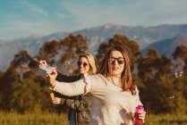 Close female friends in sunglasses blowing soap bubbles together standing on meadow in mountains — Stock Photo