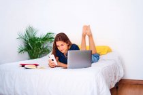 Jovem estudante deitada na cama com laptop e livros didáticos e mensagens no smartphone durante estudos on-line remotos em casa — Fotografia de Stock