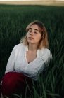 Мирная молодая женщина в белой блузке в стиле ретро сидит среди высокой зеленой травы и закрытые глаза во время отдыха в летний вечер в сельской местности — стоковое фото