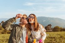 Close female friends in sunglasses blowing soap bubbles together standing in embrace on meadow in mountains — Stock Photo