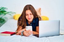 Jeune étudiante couchée sur le lit avec ordinateur portable et manuels scolaires et messagerie sur smartphone pendant des études en ligne à distance à la maison — Photo de stock