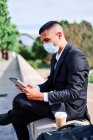 Side view of concentrated young businessman in elegant suit and protective mask reading news on tablet while having break with cup of takeaway coffee on urban street during coronavirus pandemic — Stock Photo