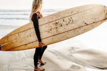 Вид збоку жінка-серфер, одягнена в гідрокостюм, стоїть озираючись з дошкою для серфінгу на пляжі під час сходу сонця на задньому плані — стокове фото