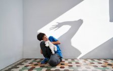 Verängstigter kleiner Junge mit Spielzeug in den Händen, der in der Nähe einer Mauer sitzt, mit dem Schatten eines wütenden gewalttätigen Elternteils mit erhobenem Arm — Stockfoto