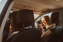 Woman with vintage camera photographing female friend driving car having trip in sunny day — Stock Photo