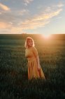 Молода жінка в старовинній сукні, дивлячись в очі, стоїть на самоті в трав'янистому полі під час заходу сонця влітку в сільській місцевості — стокове фото