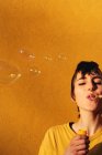 Femme moderne avec des bulles de savon perçant soufflant avec les yeux fermés à la caméra le jour ensoleillé contre le mur jaune — Photo de stock