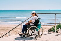 Вид збоку жінка мандрівник у інвалідному візку з рюкзаком насолоджується літньою подорожжю на пляжі біля блакитного моря, дивлячись на камеру — стокове фото