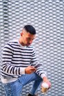 Hispanischer Mann in stylischem Outfit schaut weg und benutzt Handy, während er sich auf der Stadtstraße an die Wand lehnt — Stockfoto