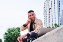 Low angle of focused young Hispanic man in stylish casual clothes working remotely on freelance project on laptop while sitting with cup of coffee near modern urban buildings — Stock Photo