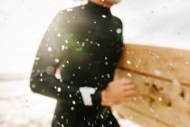 Обрізаний невпізнаваний серфінг чоловік, одягнений у гідрокостюм, біжить, дивлячись з дошкою для серфінгу на пляжі під час сходу сонця — стокове фото