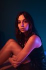 Vista laterale di tranquillo giovane modello femminile in abito seduto sul pavimento guardando la fotocamera in studio scuro con luci colorate — Foto stock