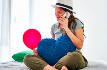 Jovem fêmea em roupas casuais e chapéu segurando balão vermelho e almofada em forma de coração navegando no smartphone enquanto celebra aniversário sozinha na cama com alimentos e suco — Fotografia de Stock
