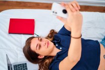 Сверху положительная молодая студентка лежит на кровати и делает селфи на смартфоне во время перерыва во время дистанционного онлайн-обучения дома — стоковое фото