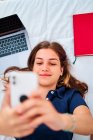 Desde arriba de la joven estudiante positiva acostada en la cama y tomando selfie en el teléfono inteligente mientras toma un descanso durante los estudios remotos en línea en casa - foto de stock