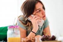 Vista lateral da jovem estudante falando no celular perto da mesa com frutas frescas e suco enquanto passa a manhã em casa — Fotografia de Stock