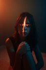 Joven modelo femenino sin emociones con proyección de luz en forma de cruz en la cara sentado en un estudio oscuro y mirando a la cámara - foto de stock