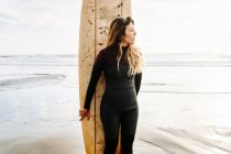 Surfer donna vestita in muta in piedi guardando lontano con la tavola da surf sulla spiaggia durante l'alba sullo sfondo — Foto stock