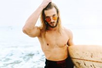 Jeune surfeur aux cheveux longs vêtu d'une combinaison et de lunettes de soleil élégantes regardant la caméra avec planche de surf vers l'eau pour attraper une vague sur la plage — Photo de stock