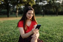Feminino em roupas de verão sentado no prado verde no parque e navegar na Internet no telefone celular enquanto se diverte no fim de semana à noite — Fotografia de Stock