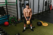 Homme athlétique avec torse nu faisant des exercices avec kettlebell lourde pendant l'entraînement actif dans le centre sportif — Photo de stock