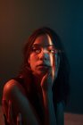 Jeune modèle féminin coûteux avec projection de lumière sur le visage assis dans un studio sombre et regardant loin — Photo de stock