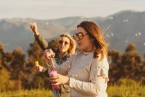 Близькі друзі-жінки в сонцезахисних окулярах дме мильні бульбашки разом, стоячи на лузі в горах — стокове фото