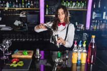 Barista femminile in abito elegante aggiungendo cubetti di ghiaccio nello shaker mentre prepara cocktail in piedi al bancone in un bar moderno — Foto stock