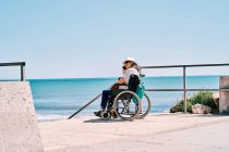 Vue latérale d'une voyageuse handicapée positive assise en fauteuil roulant et parlant sur un téléphone intelligent pendant ses vacances d'été sur la plage près de l'océan — Photo de stock