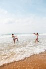 Vista lateral de amigas em trajes de banho salpicando umas às outras no oceano espumoso perto da praia de areia sob o céu azul nublado no dia ensolarado — Fotografia de Stock