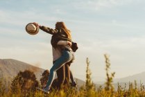 Vista lateral da mulher carregando namorada excitada nos braços girando enquanto estava no campo nas montanhas — Fotografia de Stock