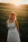 Junge Frau in weißer Schürze im Vintage-Stil, die nachdenklich wegschaut, während sie allein auf der Wiese bei Sonnenuntergang am Sommerabend auf dem Land steht — Stockfoto