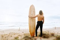 Vue de dos de surfeur méconnaissable homme aux cheveux longs habillé en combinaison debout avec planche de surf dans les dunes de sable — Photo de stock