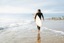 Surfer uomo vestito in muta a piedi guardando lontano con la tavola da surf verso l'acqua per catturare un'onda sulla spiaggia durante l'alba — Foto stock
