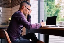 Серйозний латиноамериканський менеджер чоловічої статі у фіолетовій сорочці і окуляри друкують на клавіатурі нетбука, сидячи за вікном і працюючи над проектом у кафе. — стокове фото