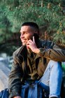 Hispanic guy in casual clothes looking away and answering phone call while standing near barrier and coniferous tree in park — Stock Photo