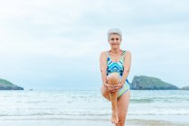 У віці сіро-волохата жінка в купальнику робить стоячи коліно підвищувати вправи на березі моря, практикуючи здоровий спосіб життя — стокове фото