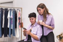 Jóvenes amigas positivas en traje de moda mirando a través de la información en el teléfono en la sala de luz con ropa en perchas - foto de stock