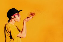 Modern female in hip hop cap and sunglasses blowing colorful confetti and having fun against yellow background — Stock Photo