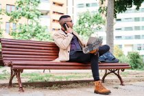 Молодий бізнесмен працює зі своїм ноутбуком на лавці парку, розмовляючи по телефону — стокове фото
