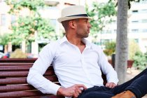Confident handsome young Hispanic male in elegant clothes and hat sitting on bench and looking away while resting on city street — Stock Photo