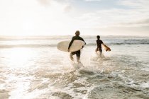 Vista posteriore di irriconoscibili amici surfisti maschili vestiti con mute che camminano con tavole da surf verso l'acqua per catturare un'onda sulla spiaggia durante l'alba — Foto stock