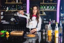 Barista donna in abito elegante aggiungendo cubetti di ghiaccio in un bicchiere mentre prepara cocktail in piedi al bancone in un bar moderno — Foto stock
