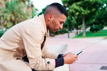 Low angle of positive well dressed young Hispanic businessman texting on smartphone and discussing news on urban street with contemporary buildings in background — Stock Photo