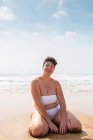 Улыбающаяся молодая женщина в купальнике, сидящая на песчаном пляже и смотрящая в камеру возле пенного океана под голубым облачным небом при дневном свете — стоковое фото