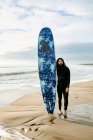 Surfista com cabelos longos e barba vestida de fato de mergulho em pé olhando para a câmera com a prancha de surf na praia durante o nascer do sol no fundo — Fotografia de Stock