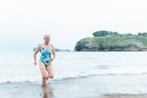 Positivo attivo in forma femminile invecchiato in costume da bagno a piedi fuori dall'acqua di mare mentre si gode la giornata estiva sulla spiaggia — Foto stock